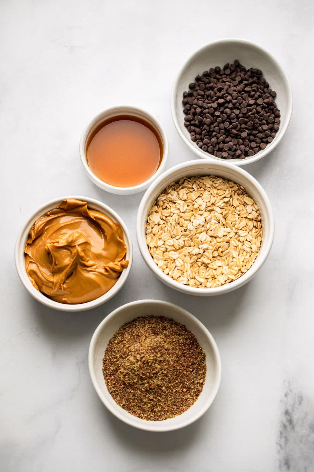 Ingredientes para bolas de energia da manteiga de amendoim em tigelas brancas individuais.