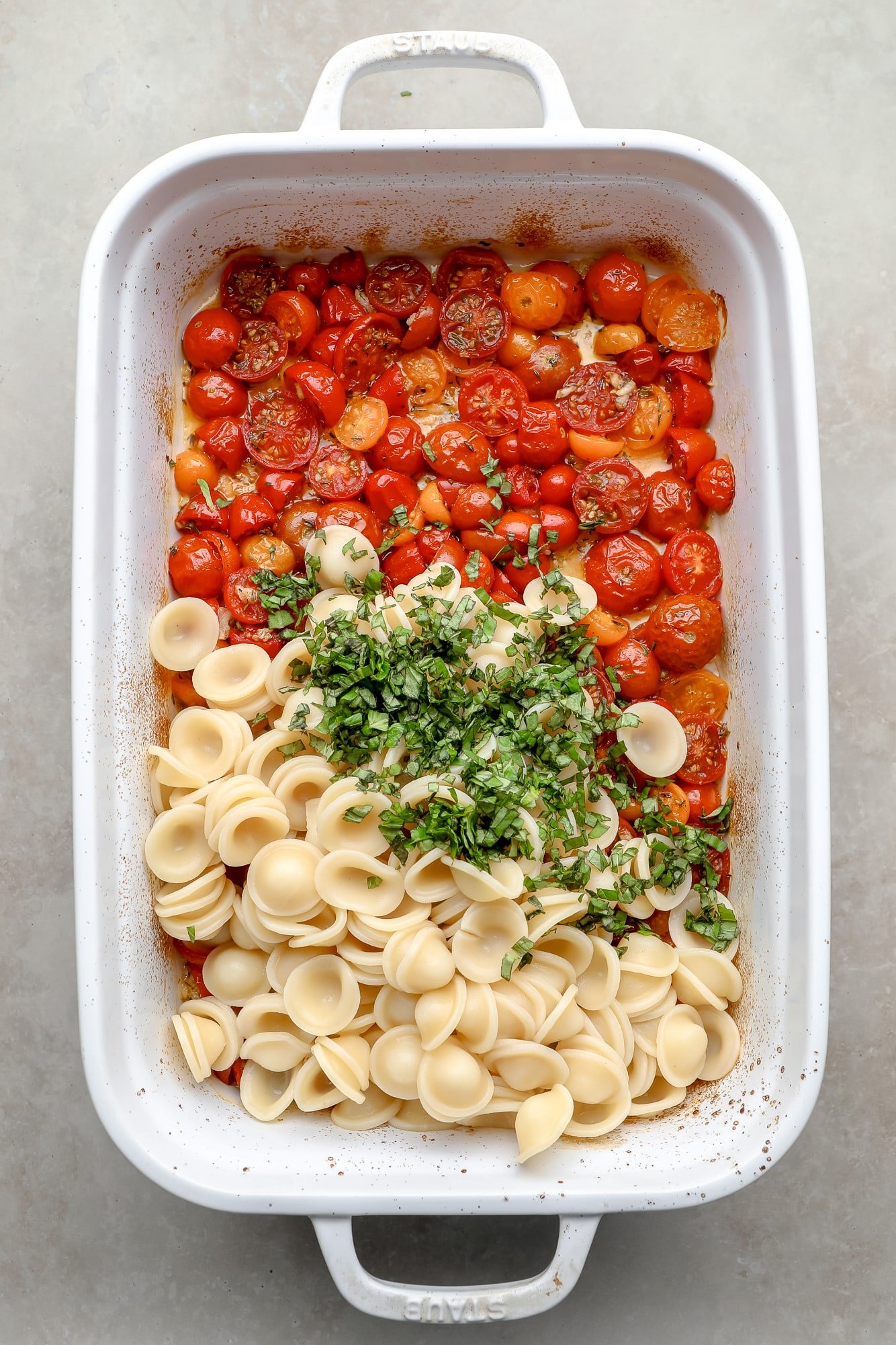 Macarrão cozido e ervas frescas em cima de tomates assados em uma assadeira branca.