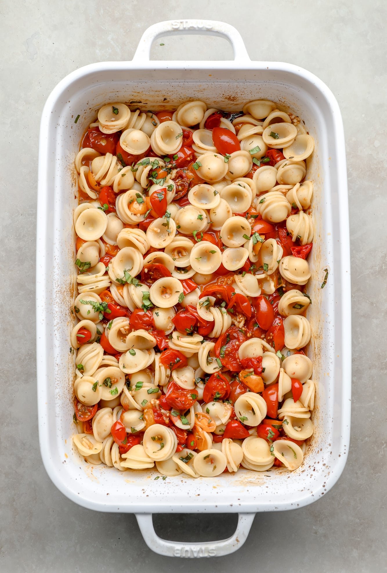 Macarrão, tomate assado e ervas jogadas juntas em uma assadeira branca.