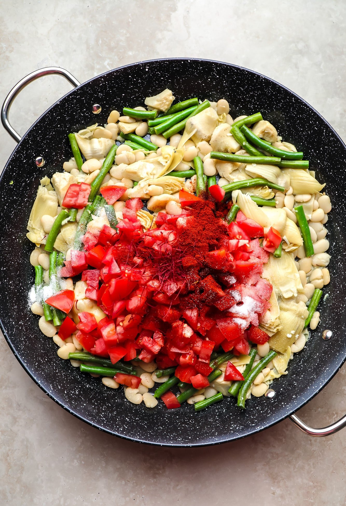Tomates picados e açafrão em cima de feijão e corações de alcachofra em uma panela preta.