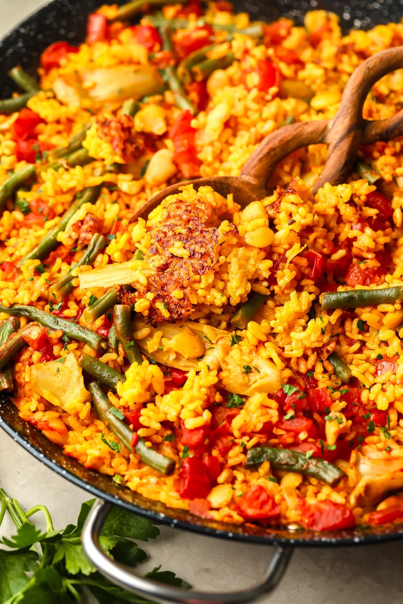 Uma colher de pau levantando uma colher de paella vegana de uma grande paella preta.